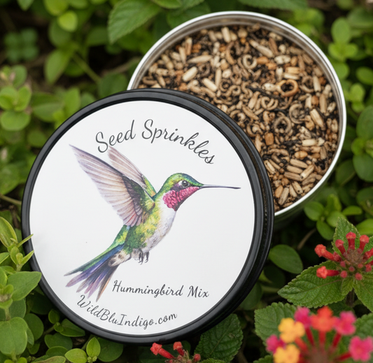 A round metal tin of seed sprinkles with the lid off, showing the mixed seed contents. The lid features a printed label with an illustration of a hummingbird and the text 'Seed Sprinkles', 'Hummingbird Mix', and 'WildBlueIndigo.